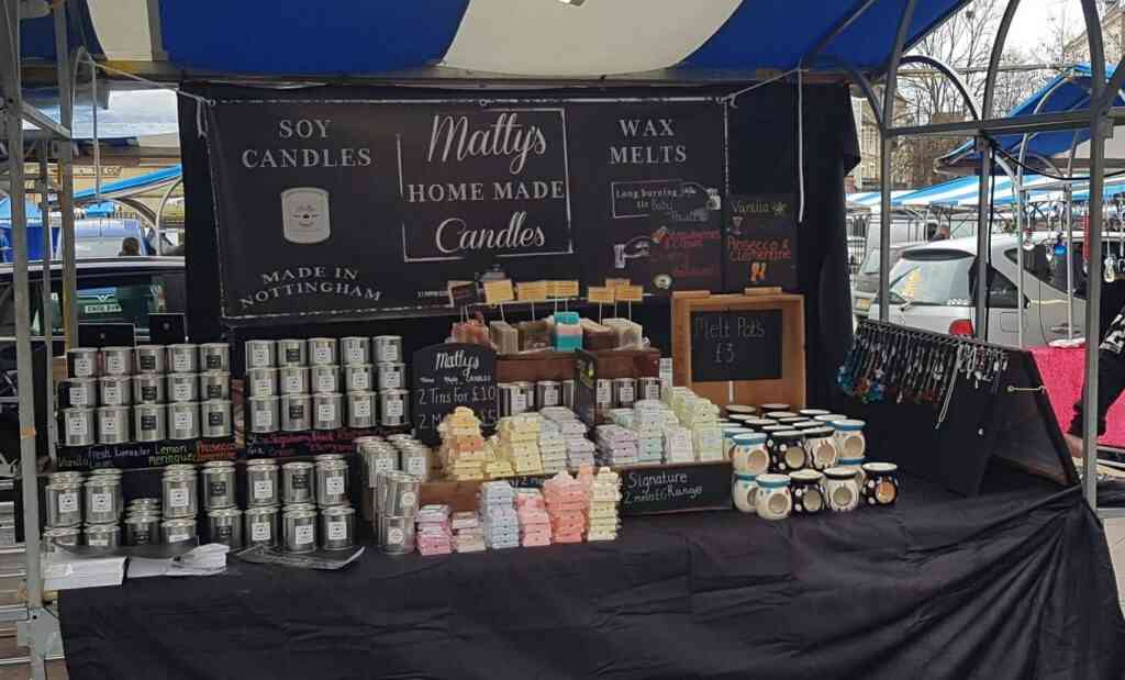 A market stall showcasing homemade candles and wax melts. The display includes an array of candles in jars, colorful wax melts, and keychains. A black banner behind the stall reads "Matty's Home Made Candles" and lists prices and product details.