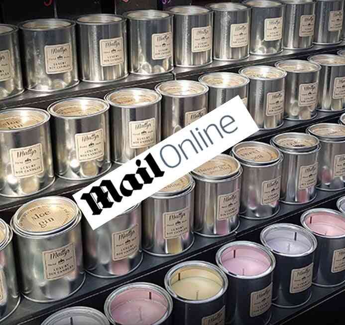 A display of multiple metal tin scented candles, some with labels that read "Melted by the Candle Alchemist." The tins by Matty's Candles are arranged on shelves, featuring unlit pink or cream-colored wax. A "Daily Mail" sign is prominently overlaid on the image.