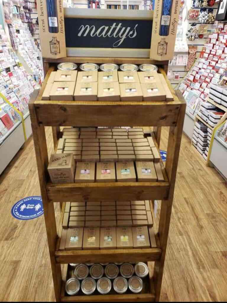 A wooden display shelf holding various candles and packaged items stands in a store aisle. The top section has a sign that reads "matty's." Stockists from Lincolnshire provide the goods. The surrounding area is filled with greeting cards and stationery items. A social distancing floor marker is visible.
