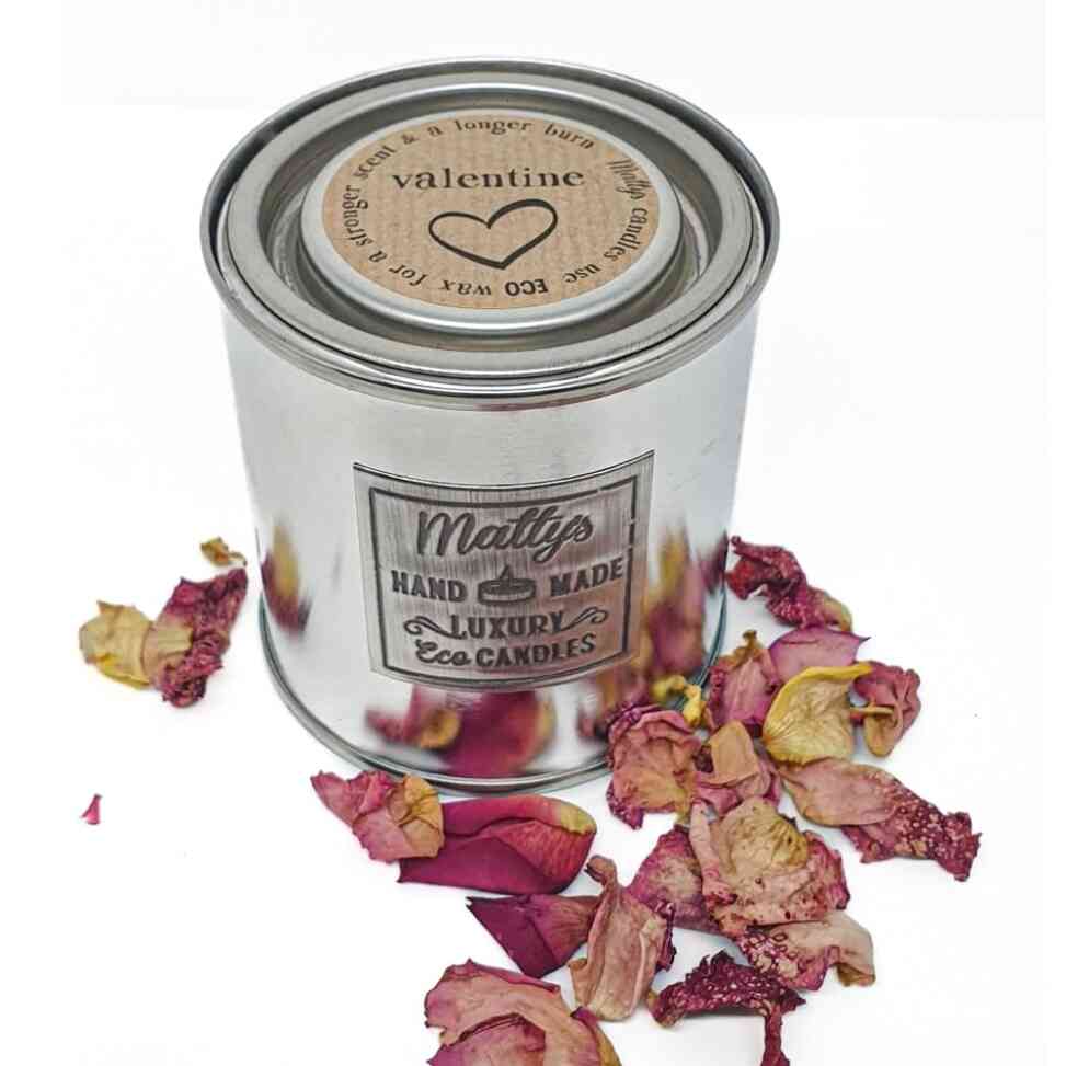 A silver tin labeled "Matty's Hand Made Luxury Eco Candles" sits surrounded by scattered dried rose petals. The lid features a circular label that reads, "valentine," with a heart symbol and text suggesting longer time use with eco wax.