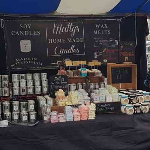 A market stall at the local fayres showcases an array of artisan soy candles and wax melts with a sign that reads "Matty's Home Made Candles Made in Nottingham." The neatly stacked candles in tins, colorful wax melts, and "Melt Pots" priced at £3 are accompanied by various labels and signs describing the products.