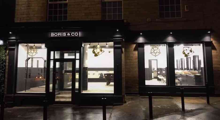 A modern barbershop named "Boris & Co" is seen at night with well-lit, large windows showcasing an interior of stylish black and white decor, barber chairs, and mirrors. The exterior features dark framing, illuminated signage, and wet pavement reflecting the lights — a true haven for grooming retailers.