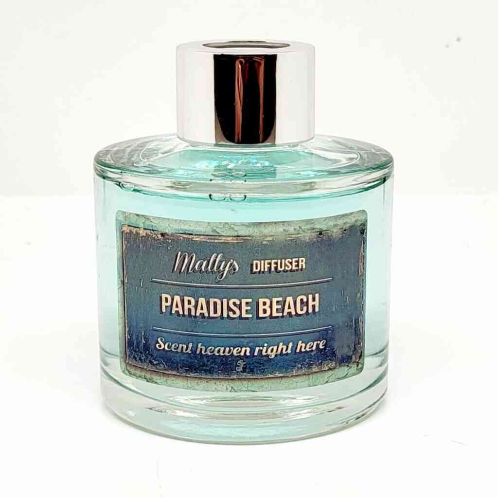 A round glass diffuser bottle filled with a light blue liquid. The bottle has a silver cap and a label that reads "Matty's Diffuser, Paradise Beach, Scent heaven right here" in blue and gold text. This elegant **Paradise Beach 90G Glass Diffuser** holds 90G of aromatic delight, capturing the essence of Paradise Beach.