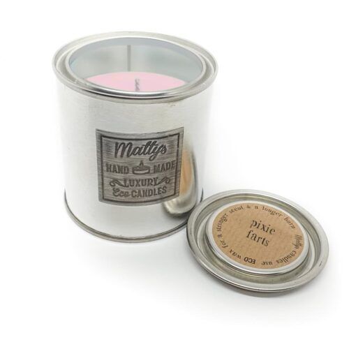 A silver cylindrical candle container labeled "Pixie Farts Candle" with an open lid sitting beside it. The pink candle inside is visible through a clear window on the top. The lid has a sticker that reads "Pixie farts.