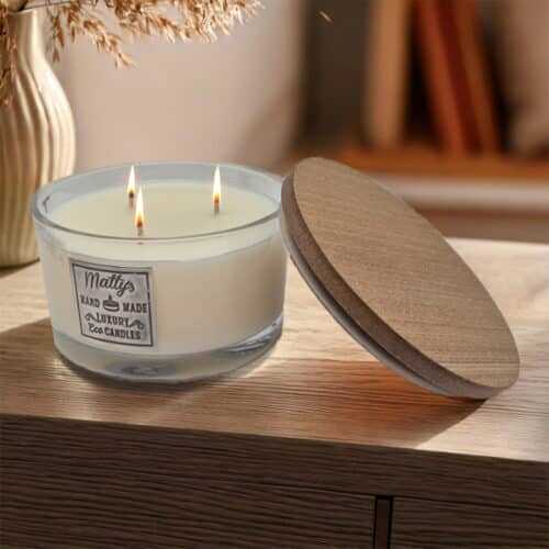 A 3-wick white candle labeled "Matty's Handmade Luxury Eco Candles" sits on a wooden surface with its wooden lid, filling the air with the refreshing scent of cucumber spa as soft sunlight glows in the background.